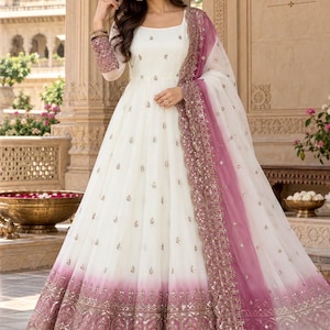 May include: A floor-length white gown featuring a square neckline, long sleeves with embellished cuffs, and a flared skirt with a pink ombre hem. The dress is adorned with small gold floral motifs and a wide, intricate metallic border at the base.