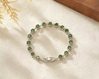 May include: A silver-toned bracelet featuring a series of round, faceted green stones set in individual metal bezels. The bracelet is connected by small circular links and includes a secure metal clasp mechanism at the center.