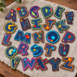 Puede incluir: Una colección de veintiséis letras del alfabeto hechas de tela de mezclilla, cada una con bordes bordados coloridos y costuras decorativas en varios patrones, formas y colores brillantes, dispuestas sobre una superficie de tela texturizada.