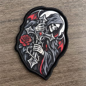 Puede incluir: Un parche de tela que muestra una figura de la parca con una capa oscura con capucha y alas rojas, sosteniendo una cruz plateada envuelta en espinas. Una rosa roja se sitúa a la izquierda de la cruz, sobre un fondo circular blanco.