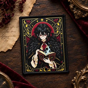 Puede incluir: Un parche bordado rectangular que muestra la ilustración de una persona con cabello negro y gafas, vistiendo una prenda de plumas oscuras, sosteniendo un libro abierto con llamas azules que surgen de las páginas, enmarcado por alas doradas.