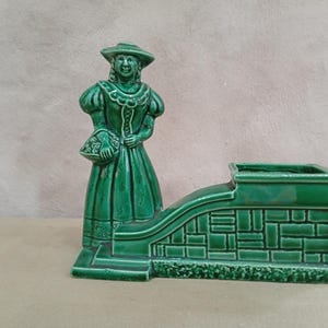 May include: A glossy emerald green ceramic planter featuring a standing figure wearing a wide-brimmed hat and a long dress, holding a basket. The figure is attached to a rectangular, brick-patterned basin with a curved side wall and base.