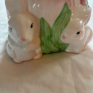 May include: A ceramic planter shaped like a large pink tulip flower with two white rabbits sculpted on the front. The rabbits are positioned on either side of green leaves, featuring painted pink noses and dark eyes against a glossy finish.