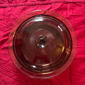 Puede incluir: Una tapa de cristal redonda de color ámbar con un pomo central. La tapa tiene una superficie lisa y transparente con un patrón decorativo de rayos de sol grabado en el cristal alrededor del pomo central, vista desde arriba.