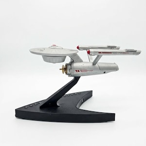 May include: A silver-colored model of the Starship Enterprise NCC-1701, featuring a saucer section, a secondary hull, and two nacelles. The model is mounted on a black, V-shaped display stand with five circular buttons along the base.
