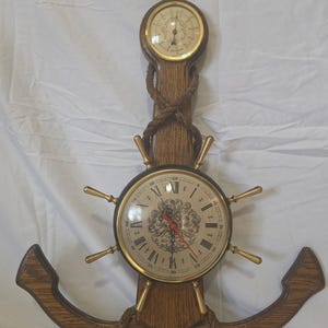 May include: A wall-mounted nautical clock and barometer set shaped like an anchor. The wooden anchor frame features a ship's wheel clock with Roman numerals and gold spokes, topped by a circular barometer. A decorative rope is wrapped around the frame.