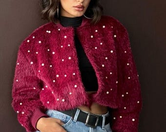 May include: A cropped, deep burgundy fuzzy jacket featuring a round neckline, long sleeves with ribbed cuffs, and scattered white faux pearl embellishments throughout the front and sleeves. The jacket is worn over a black top with light blue jeans.