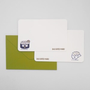 May include: A stationery set featuring a green envelope, two white notecards, and a design of a sushi character. The notecards have the text "BAD BADTZ-MARU" and a copyright notice. The sushi character is in blue and brown.