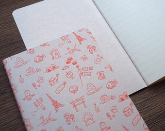Puede incluir: Cuaderno abierto con una cubierta color crema con un patrón repetitivo de ilustraciones japonesas rojas. Las páginas abiertas revelan papel rayado. El cuaderno está sobre una superficie de madera.
