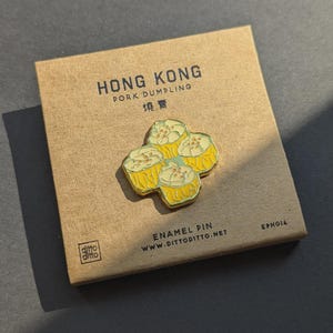 May include: A yellow enamel pin depicting four dim sum dumplings, displayed on a brown card. The card reads "HONG KONG PORK DUMPLING" and includes Chinese characters. A food-themed accessory.