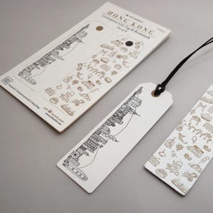 Könnte beinhalten: Off-white Letterpress-Geschenkanhänger und -Lesezeichen mit schwarzen und goldenen Illustrationen. Die Anhänger und Lesezeichen zeigen ein Stadtbilddesign und kleine Symbole. Der Text "HONG KONG Letterpress Gift Tag & Bookmark" ist sichtbar.