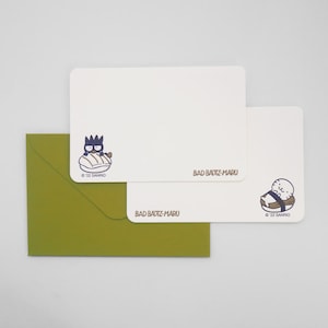 May include: A stationery set featuring Bad Badtz-Maru designs. The set includes white notecards with Bad Badtz-Maru illustrations and text, along with a green envelope. The text on the cards reads "BAD BADTZ-MARU" and "© 22 Sanrio".