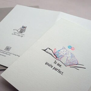 May include: A folded greeting card with a cat illustration. The front of the card has the text "To me, you're perfect." The inside of the card has a cat wearing glasses and the text "Inspired by movies Love Actually".