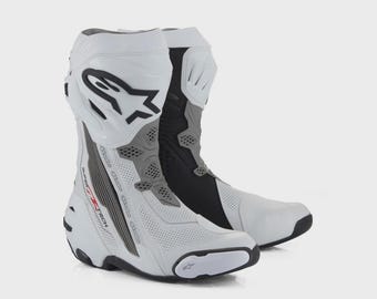 Puede incluir: Un par de botas de carreras de moto blancas, grises y negras con paneles de cuero perforado, una estructura rígida de soporte para el tobillo y el logo de Alpinestars en la espinilla. El lateral muestra el texto Supertech en letras rojas y negras.