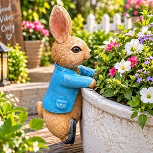 Op de afbeelding: Een bruin konijnenbeeldje met een blauw jasje, gepositioneerd alsof het over de rand van een grote, witte, ronde plantenbak vol witte, roze en paarse bloemen leunt. Het konijn heeft grote oren, donkere ogen en een textuur van vacht.