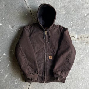 Könnte beinhalten: Eine dunkelbraune Kapuzenjacke mit durchgehendem Frontreißverschluss, gerippten Bündchen und einem gerippten Saum. Die Jacke verfügt über ein schwarzes Innenfutter, eine Kapuze mit Kordelzug und ein kleines quadratisches Logo-Patch.