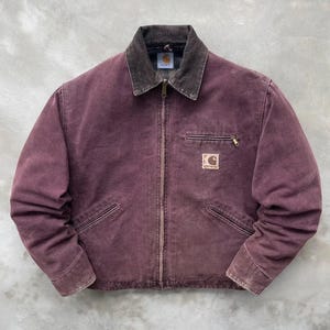 May include: A faded plum-coloured canvas jacket featuring a dark brown corduroy collar, a full-length front zip, and two slanted lower pockets. A small zipped chest pocket is present, along with a square Carhartt brand patch on the chest.