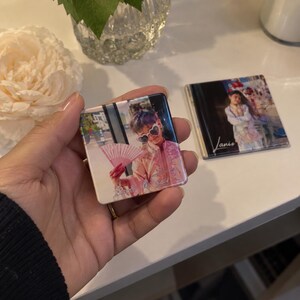Peut inclure: Deux miroirs de poche carrés avec des impressions photographiques. L'un montre une personne portant des lunettes de soleil en forme de cœur et tenant un éventail rose. L'autre affiche une personne en tenue blanche avec le nom Janis écrit en blanc.