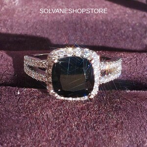 Op de afbeelding: Een ring met een grote, donkere, kussengeslepen middensteen omringd door een halo van kleine, heldere, fonkelende steentjes. De ringband is gespleten en ingelegd met extra heldere steentjes. De tekst SOLVANESHOPSTORE is bovenaan zichtbaar.