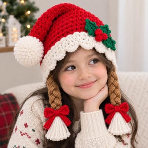 Könnte beinhalten: Eine rot-weiße Häkel-Weihnachtsmütze mit einem großen weißen Bommel, einem gewellten weißen Rand und einer Applikation aus grünen Stechpalmenblättern und roten Beeren. Daran befestigt sind zwei dicke, hellbraune Zöpfe mit roten Schleifen.