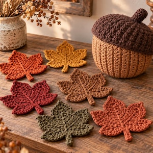 May include: A collection of six crocheted leaf coasters in shades of orange, yellow, brown, red, and olive green, accompanied by a large, round, brown crocheted acorn-shaped container with a textured cap, all arranged on a wooden surface.
