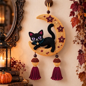 Peut inclure: Une décoration murale au crochet représentant un chat noir aux yeux violets avec un petit grelot doré, perché sur un croissant de lune jaune orné d'étoiles rouge foncé. Deux ornements en forme de gland avec de longs pompons rouge foncé pendent.