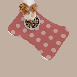 Op de afbeelding: Een botvormige voermat voor huisdieren in oudroze met een lichtroze stippenpatroon. Een kleine hond met wit en bruin haar eet uit een grijze kom gevuld met droge brokken die op de mat staat. De tekst CatsNDogsCo staat onderaan.
