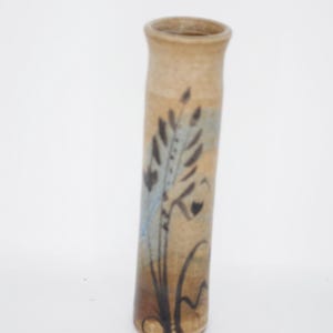 May include: A tall, cylindrical ceramic vase with a tan, speckled base colour. The exterior features a hand-painted botanical design in dark brown and muted blue tones, depicting slender stalks of wheat or grass rising from the base to the rim.