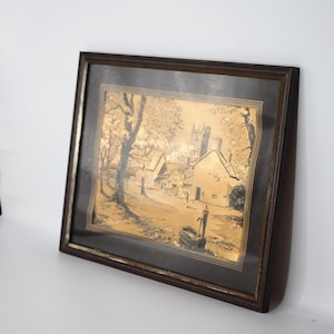May include: A rectangular framed wall art piece featuring a metallic gold-toned etching of a village scene with trees, houses, a church tower, and a water pump. The artwork is encased in a dark brown wooden frame with a black matting border.