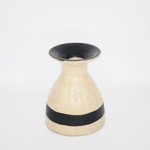 May include: A small, cream-coloured ceramic vase with a speckled texture. The vase features a wide, flared rim painted in solid black and a single, thick horizontal black band encircling the lower portion of the body, creating a contrast.