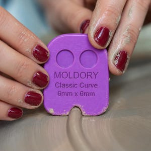 Pode incluir: Uma ferramenta de cerâmica roxa Moldory Classic Curve com dois recortes circulares no topo e o texto "MOLDORY Classic Curve 6mm x 6mm" em relevo na frente. A ferramenta possui um entalhe curvo na borda inferior para moldar a argila.
