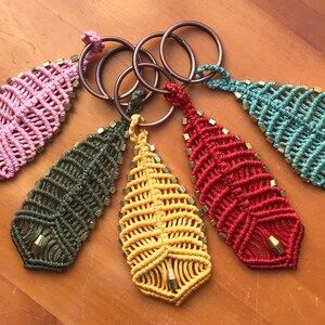 Puede incluir: Cinco llaveros de plumas de macramé de colores vivos con detalles dorados. Cada llavero está unido a un anillo de metal.