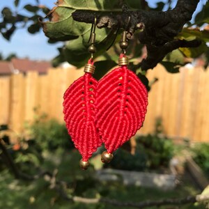 Op de afbeelding: Een paar rode, bladvormige oorbellen gemaakt met macramé en gouden metalen accenten.