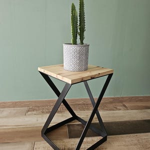Peut inclure: Une table d'appoint carrée avec un plateau en bois naturel clair et un cadre en métal noir doté d'une base en forme de X. Un grand cactus vert est planté dans un pot cylindrique gris avec un motif géométrique blanc en relief.