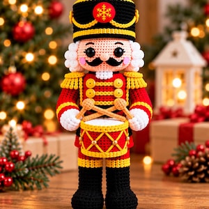 Puede incluir: Un muñeco cascanueces de ganchillo con un sombrero negro y amarillo con un emblema de copo de nieve rojo, una chaqueta roja con botones amarillos y botas negras. La figura sostiene dos baquetas de madera sobre un tambor rojo y amarillo.