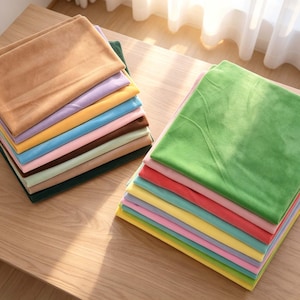 May include: Two stacks of folded fabric squares in a variety of solid colors including tan, lavender, yellow, blue, pink, brown, light green, dark green, and bright green. The fabric has a soft, matte texture and is neatly layered in two piles.