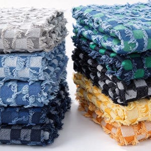 May include: Two stacks of folded square napkins with a frayed edge finish. The left stack features shades of gray and blue in a checkered pattern. The right stack displays a vibrant checkered pattern in blue, green, black, yellow, and orange.