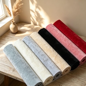 Può includere: Una collezione di sei tappetini in tessuto arrotolati in una sfumatura di colori che include grigio chiaro, crema, grigio medio, beige, nero, rosa antico e rosso intenso, disposti in fila diagonale su una superficie in legno accanto a un vaso beige.