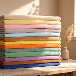 May include: A stack of folded rectangular towels in a variety of solid colors including beige, yellow, pink, light blue, brown, orange, green, purple, and dark blue, arranged in a vertical pile with a soft, plush, and textured fabric surface.