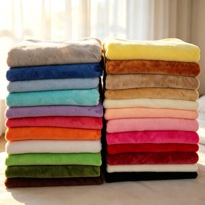 Könnte beinhalten: Zwei Stapel gefalteter Plüschdecken in einer Vielzahl von Unifarben, darunter Gelb, Blau, Rosa, Grün, Rot, Orange, Lila, Braun, Schwarz und Weiß. Jede Decke hat eine weiche, flauschige Textur und ist ordentlich zu einem Rechteck gefaltet.
