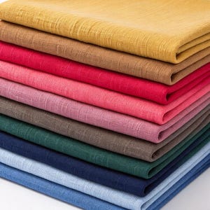 May include: A stack of folded fabric pieces in various colours including mustard yellow, tan, red, pink, mauve, brown, dark green, navy blue, light blue, and medium blue. Each piece of fabric features a visible, textured, cross-hatched weave pattern.