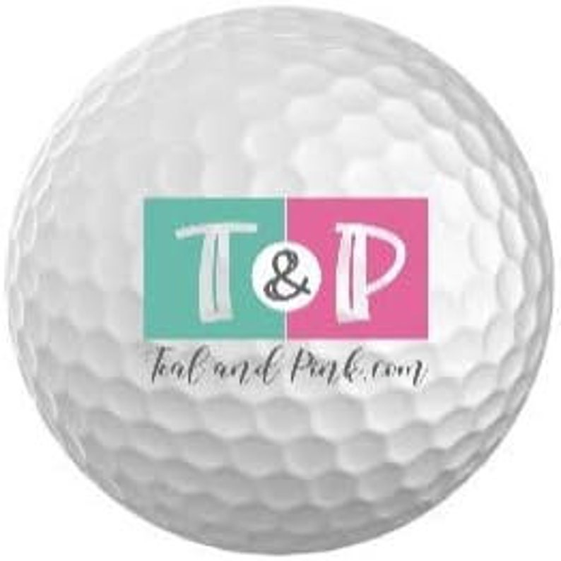 Puede incluir: Bola de golf blanca con un logotipo turquesa y rosa que dice "T&P" y "teal and pink.com"
