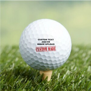 Puede incluir: Una bola de golf blanca con texto negro que dice "CUSTOM TEXT AND/OR IMAGE UPLOAD" y un sello rojo que dice "CUSTOM MADE".