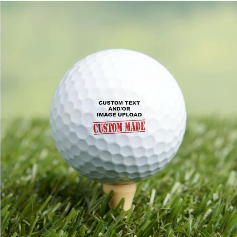 Puede incluir: Una bola de golf blanca con texto negro que dice "CUSTOM TEXT AND/OR IMAGE UPLOAD" y un sello rojo que dice "CUSTOM MADE".