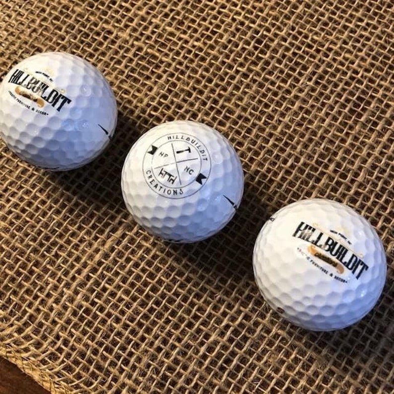 Puede incluir: Tres bolas de golf blancas con letras negras. La primera bola dice "Hill Build It" con un peque&ntilde;o logotipo de una casa. La segunda bola dice "Hill Build It" con un logotipo de br&uacute;jula y las palabras "HTH Creations". La tercera bola dice "Hill Build It" con un peque&ntilde;o logotipo de una casa.