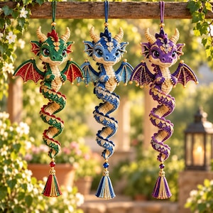 May include: Three crocheted dragon ornaments hanging vertically. The dragons are green, blue, and purple, each featuring large eyes, horns, wings, and a long coiled tail ending in a decorative tassel with a gold bead and a colored crystal.