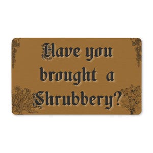 May include: A rectangular brown doormat featuring the black text Have you brought a Shrubbery? in a gothic font. The design includes detailed black botanical illustrations of plants and flowers in the corners, providing a decorative border.