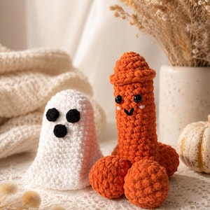May include: A white crocheted ghost figure with three black circular felt eyes and mouth, positioned next to an orange crocheted phallic-shaped figure with black plastic safety eyes and a stitched smile, both crafted from soft yarn.