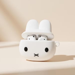 Peut inclure: Un étui de protection en silicone blanc pour écouteurs sans fil, en forme de lapin avec deux oreilles dressées sur le couvercle. L'avant présente deux yeux noirs circulaires et une bouche en forme de croix noire. Deux écouteurs blancs sont visibles.