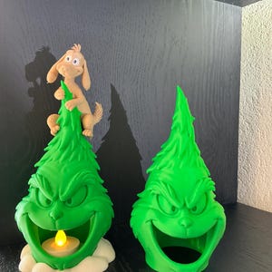 Op de afbeelding: Twee felgroene, kegelvormige decoratieve figuren met een gebeeldhouwd, grijnzend gezicht en gefronste wenkbrauwen. Eén figuur is bekroond met een beige hondje en bevat een klein, geel oplichtend LED-theelichtje in de open mond.